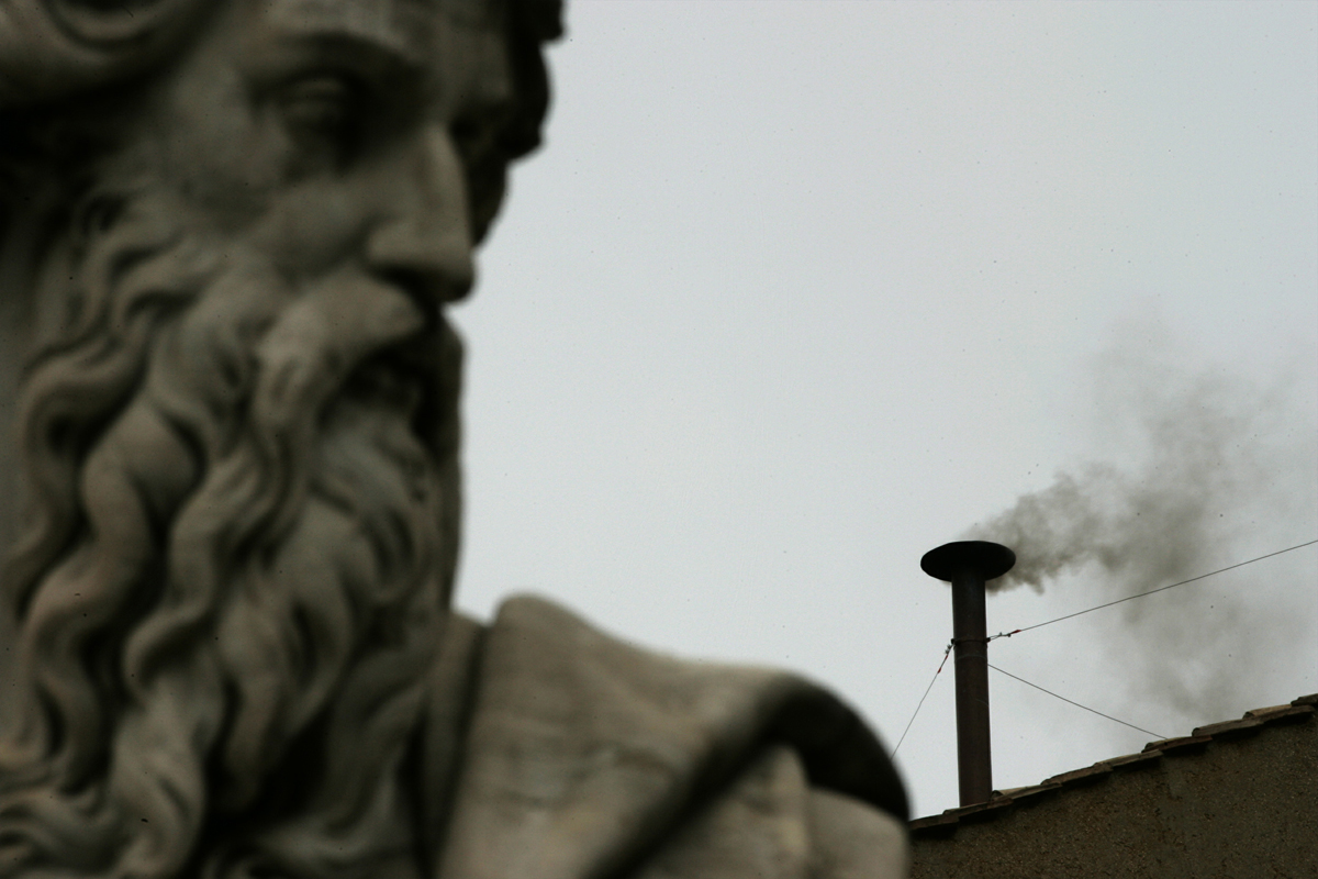 A statue looks down on black smoke rising from the chimney above the Sistine Chapel in the Vatican, ..